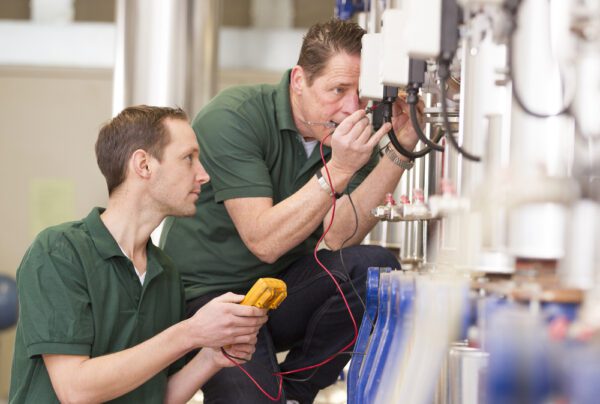 Two technicians use a voltmeter to check the calibration of production equipment as part of MRO.