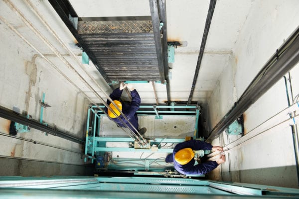 Two mechanics at work fixing adjusting elevator hoist in response to a maintenance request.