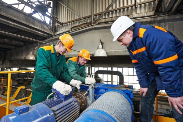 A middle-aged maintenance worker supervises two younger technicians as they repair a winch motor with a wrench.