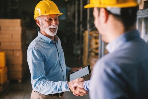 Two maintenance workers in hard hats shaking hands, one holding a tablet, in a warehouse, representing how good maintenance managers can positively impact the team.