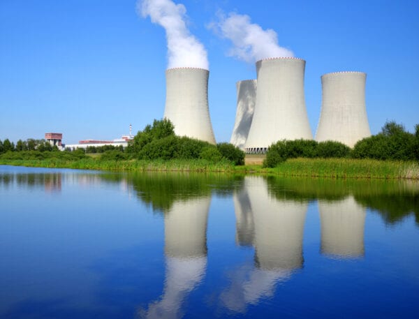 Power plant smoke stacks emitting smoke against a blue sky with trees and water nearby, referring to power plant maintenance.