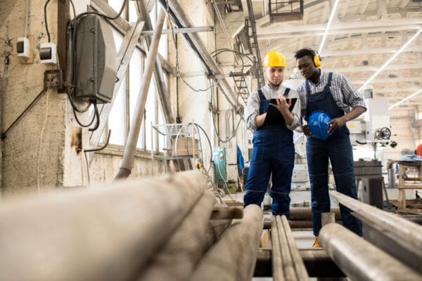 Maintenance technicians surrounded by pipes with hardhats on floor discussing maintenance efficiency killers using a tablet.