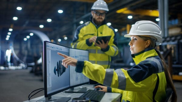 Man with tablet and woman on desktop pointing to screen with hard hats, managing maintenance work.