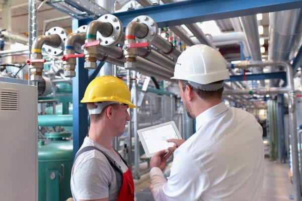 Two young male technicians looking at asset data on a tablet near some piping to help identify whether the asset is critical and should be included in the organization’s preventive maintenance program.