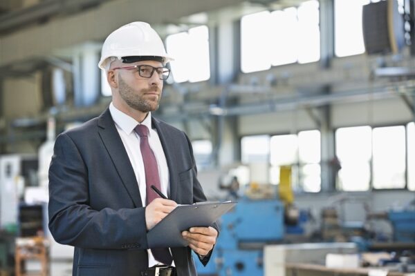 Middle-aged man in suit and hard hat writing notes on a clipboard while on factory floor