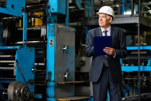 Executive in hard hat with clipboard standing near machinery.