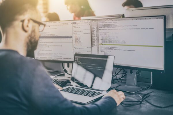 Software engineer sitting at double monitor desktop with laptop performing software validation testing.