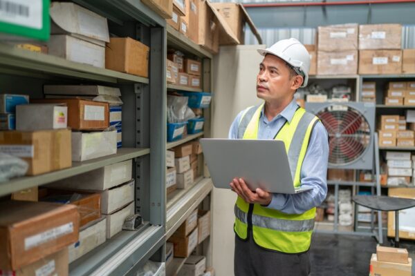 Man holding laptop in an inventory storeroom, tracking inventory with CMMS software.
