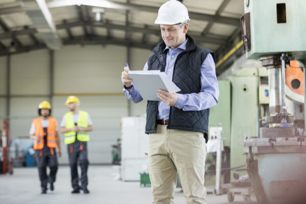 Medium shot of a middle-aged maintenance manager reviewing paper work orders on a factory floor.